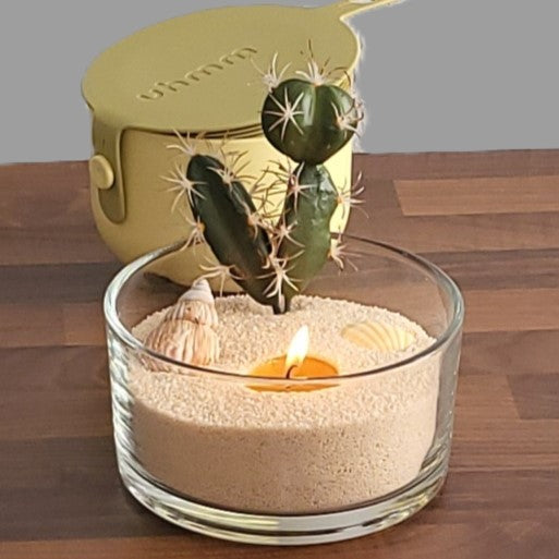 Kleine Glasschale befüllt mit beigem Kerzensand, dekoriert mit 2 Muscheln und einem Sukkulenten Imitat zum stecken. Im Hintergrund eine kleine 0,7 L Schüssel von uhmm in gelb mit Deckel in olive.